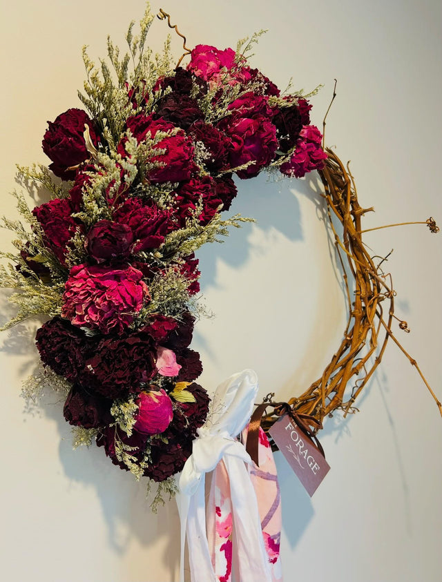 A dried peony wreath by The Forage, deep red and pink blooms on a twisted vine ring with a ribbon